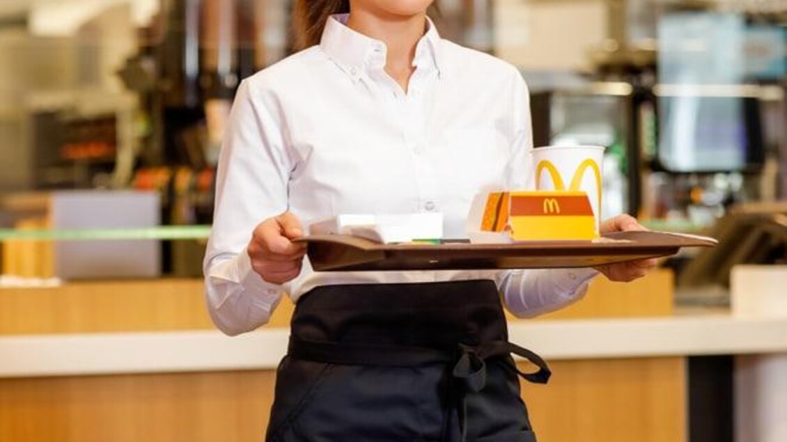 Eine Mitarbeiterin hält ein Tablett mit Essen in einem Restaurant. Im Hintergrund sind Küchenbereich und Burger-Menü sichtbar. | © mcdonalds-oesterreich