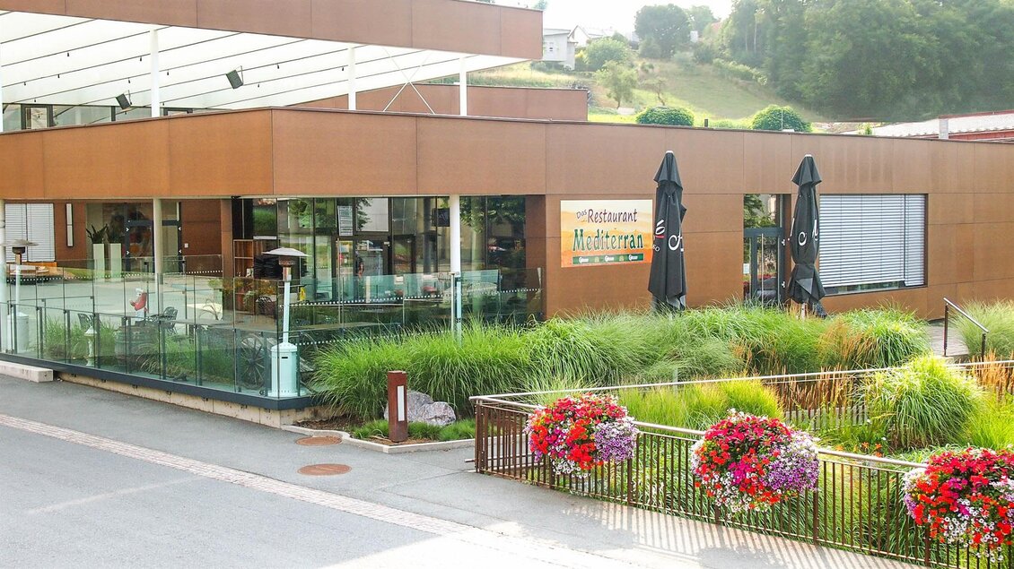 Ein modernes Restaurant mit großen Glasfenstern und einer einladenden Terrasse. Bunte Blumen und Sonnenschirme schmücken den Außenbereich. | © Mediterran - Rathaus Restaurant