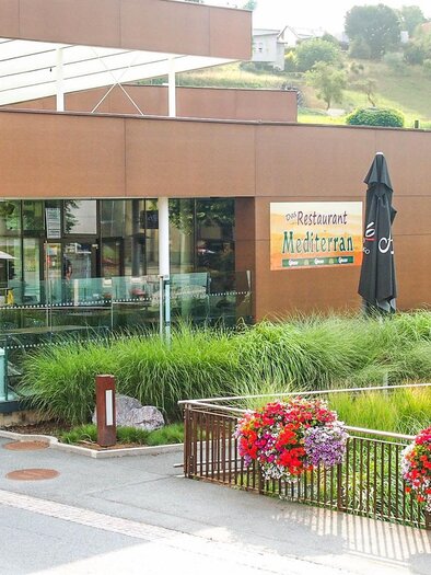 Ein modernes Restaurant mit großen Glasfenstern und einer einladenden Terrasse. Bunte Blumen und Sonnenschirme schmücken den Außenbereich. | © Mediterran - Rathaus Restaurant