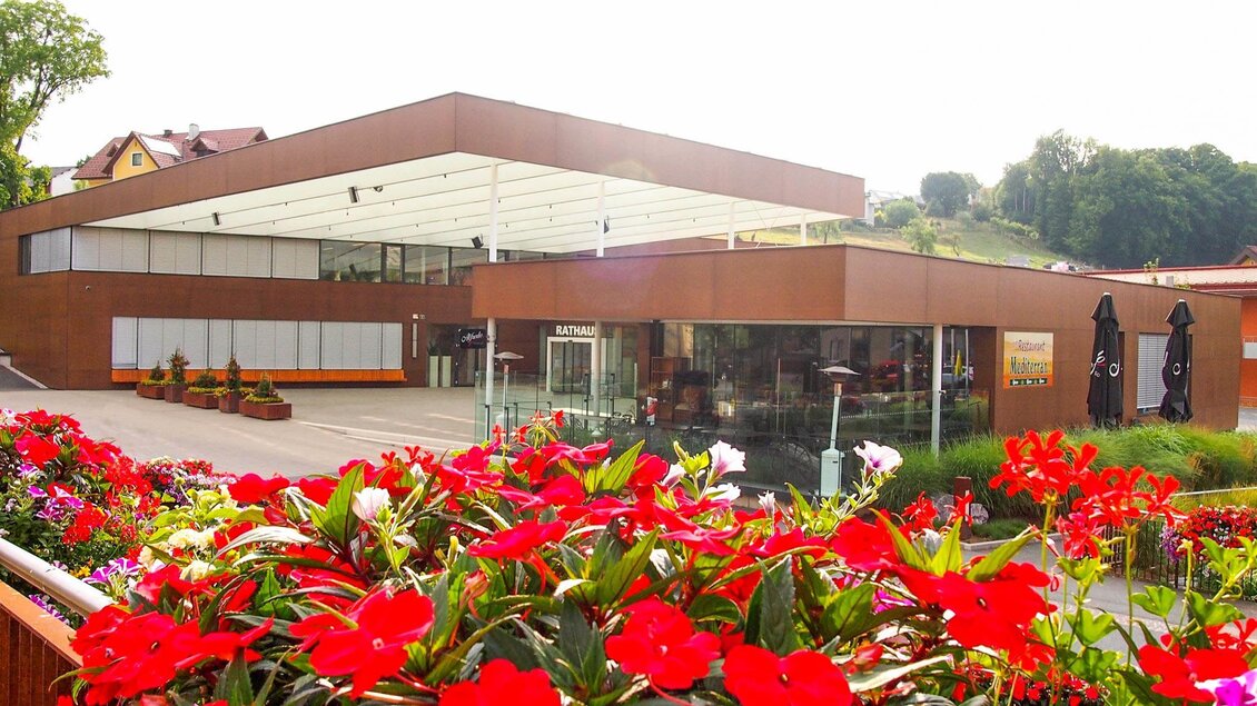 Ein modernes Gebäude mit großen Fenstern und einem einladenden Restaurant. Im Vordergrund blühen bunte Blumen, die eine freundliche Atmosphäre schaffen. | © Mediterran - Rathaus Restaurant