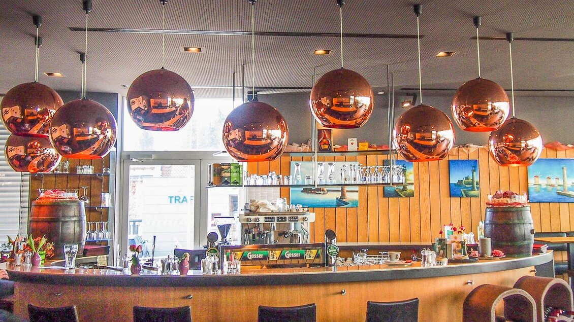 Eine moderne Bar mit einer langen Theke und stilvollen Kupferlampen hängt darüber. Im Hintergrund sind Bilder von Landschaften zu sehen, die eine einladende Atmosphäre schaffen. | © Mediterran - Rathaus Restaurant