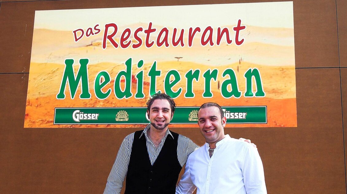Zwei Männer stehen vor einem großen Schild, auf dem „Das Restaurant Mediterran“ steht. Der Hintergrund erinnert an eine Wüstenlandschaft. | © Mediterran - Rathaus Restaurant
