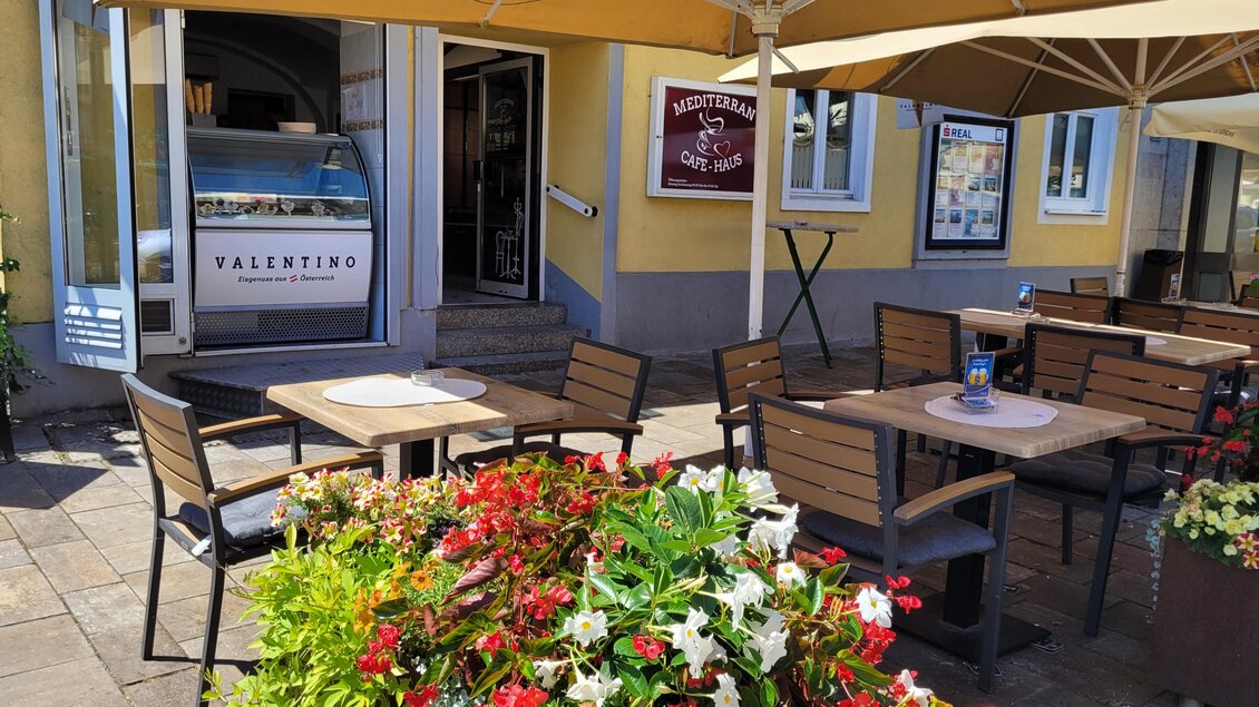 Ein gemütlicher Außenbereich mit Tischen und Stühlen, umgeben von blühenden Pflanzen. Im Hintergrund ist ein Restaurant mit einem einladenden Eingang zu sehen. | © TV Südsteiermark - Irene Löschnig