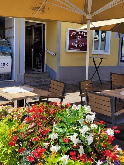 A cozy outdoor area with tables and chairs, surrounded by blooming plants. In the background, a restaurant with an inviting entrance can be seen. | © TV Südsteiermark - Irene Löschnig
