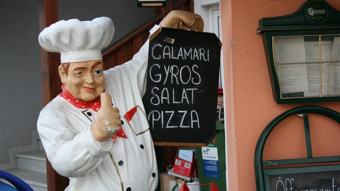 Ein freundlicher Koch mit einer Schürze zeigt ein Schild mit Speisen. Die angebotenen Gerichte umfassen Calamari, Gyros, Salat und Pizza. | © Schilcherland Steiermark