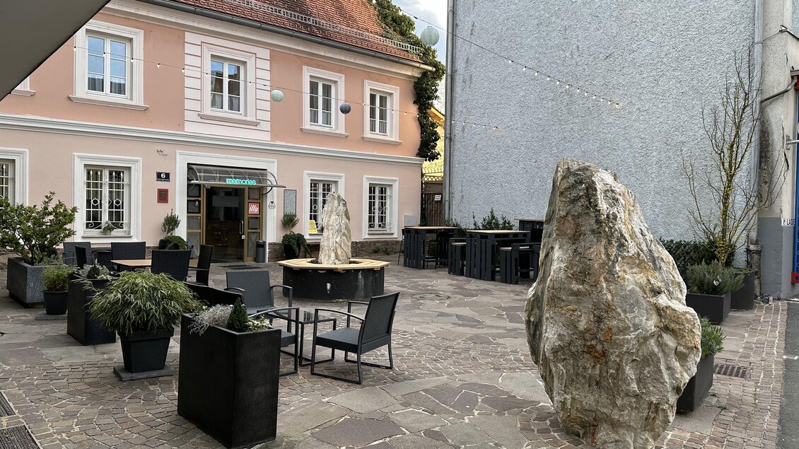 Ein gemütlicher Innenhof mit Terrassenmöbeln und einem großen Stein. Im Hintergrund steht ein charmantes Gebäude mit rosa Fassade. | © Café & Bar Memories – Julia Bernsteiner