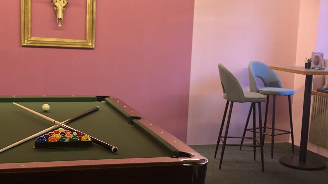Ein Billardtisch mit Kugeln in einem Raum mit rosa Wänden. Im Hintergrund steht ein Tisch mit Stühlen. | © Café & Bar Memories – Julia Bernsteiner