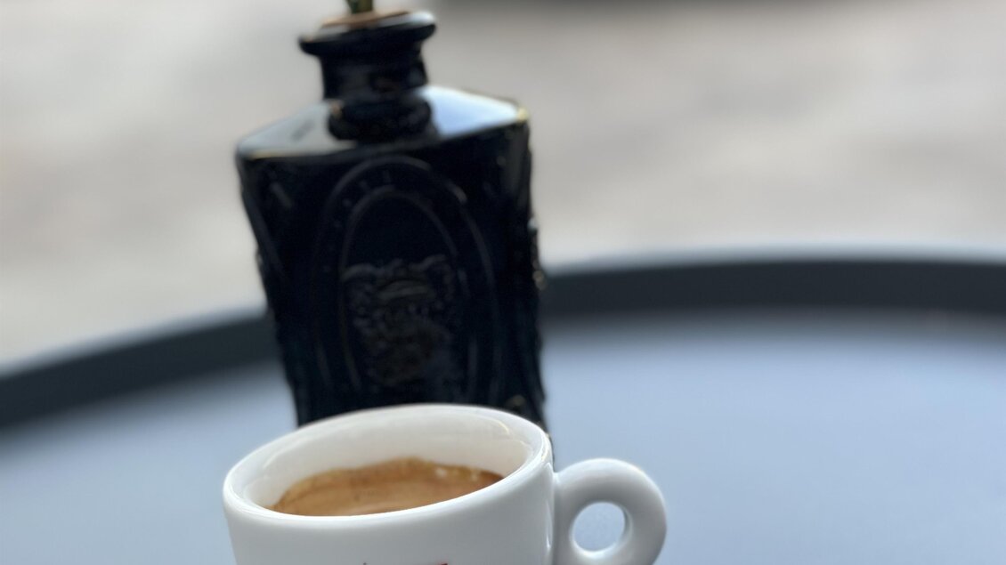 Eine Tasse Kaffee steht auf einem Tisch neben einer eleganten Vase mit einer Blume. Der Tisch ist schlicht und der Hintergrund ist verschwommen. | © Café & Bar Memories – Julia Bernsteiner