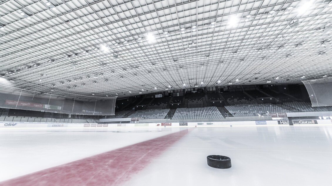 Auf der glatten Eisfläche im Merkur Eisstadion liegt ein Puck auf dem Eis. Die helle Decke und die umliegenden Tribünen sind deutlich sichtbar. | © MCG - Krug