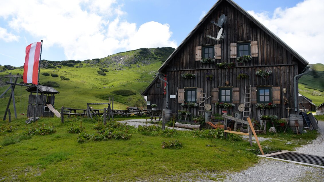 Eine traditionelle Berghütte aus Holz in einer grünen Landschaft. Im Hintergrund sind sanfte Hügel und ein klarer Himmel zu sehen. | © Naturpark Mürzer Oberland