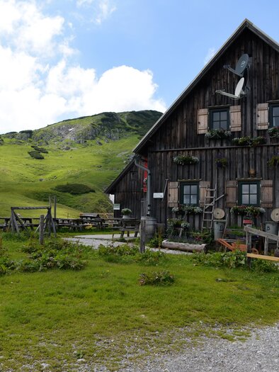 A traditional wooden mountain cabin in a green landscape. In the background, gentle hills and a clear sky can be seen. | © Naturpark Mürzer Oberland
