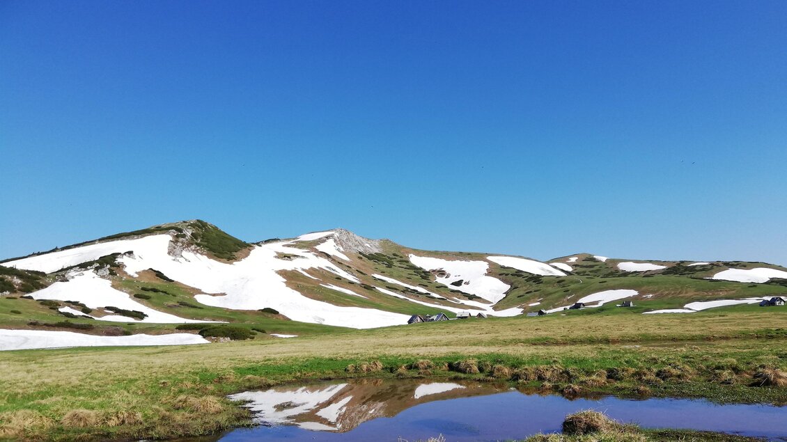 Eine grüne Landschaft mit schneebedeckten Bergen im Hintergrund. Der klare blaue Himmel spiegelt sich in einer kleinen Wasserstelle. | © Naturpark Mürzer Oberland