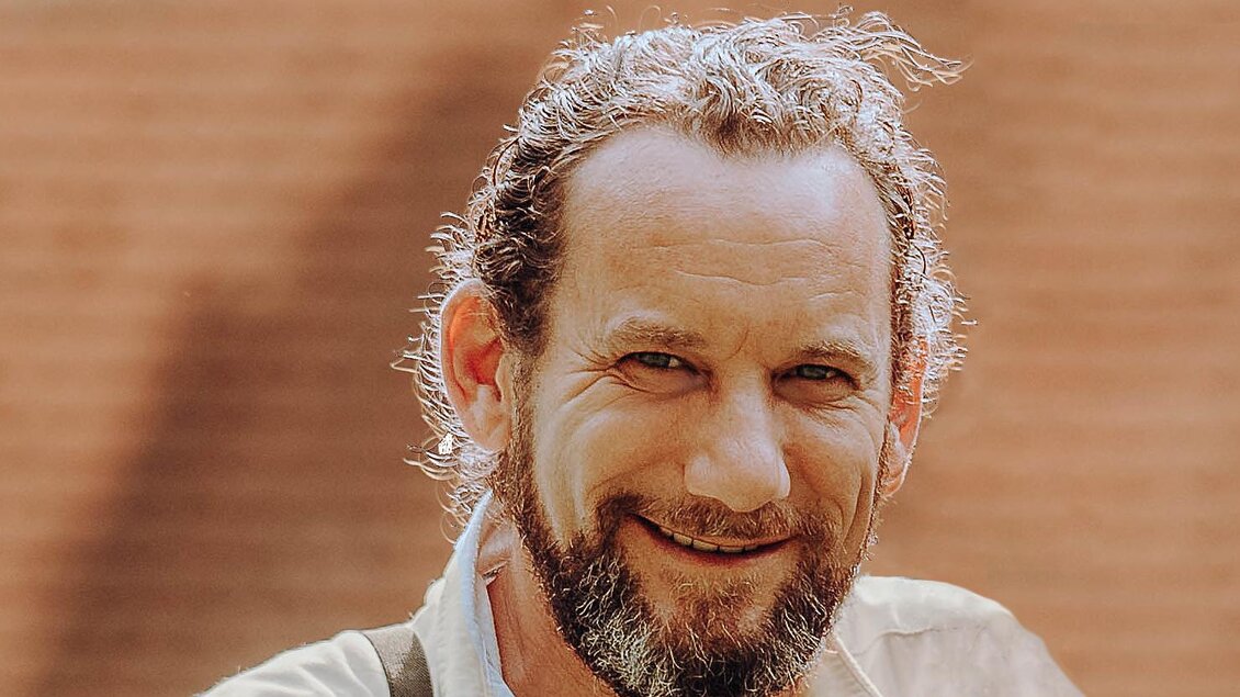 Ein Paar steht lächelnd auf einer Wiese, umgeben von Bäumen und einem historischen Gebäude im Hintergrund. Die Atmosphäre wirkt freundlich und romantisch. | © GenussReich