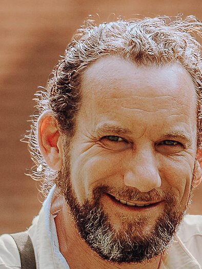 A couple is smiling on a meadow, surrounded by trees and a historic building in the background. The atmosphere feels friendly and romantic. | © GenussReich