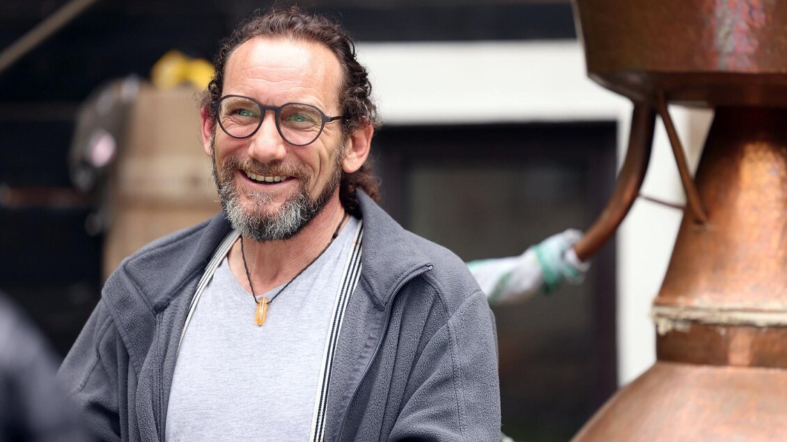 Ein lächelnder Mann mit Brille steht vor einer Kupferbrennerei. Im Hintergrund sind unscharf weitere Geräte zu erkennen. | © Mike Shane