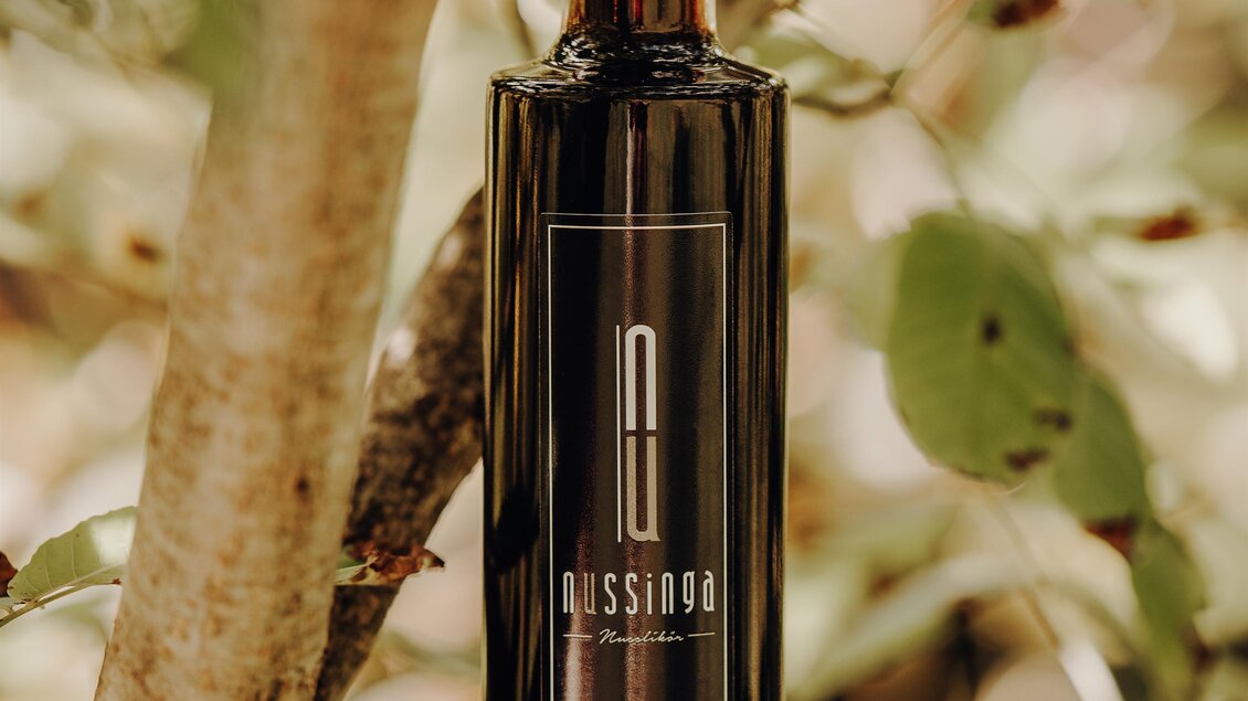 Eine elegante Flasche Öl steht auf einem Baumast. Im Hintergrund sind grüne Blätter sichtbar. | © GenussReich