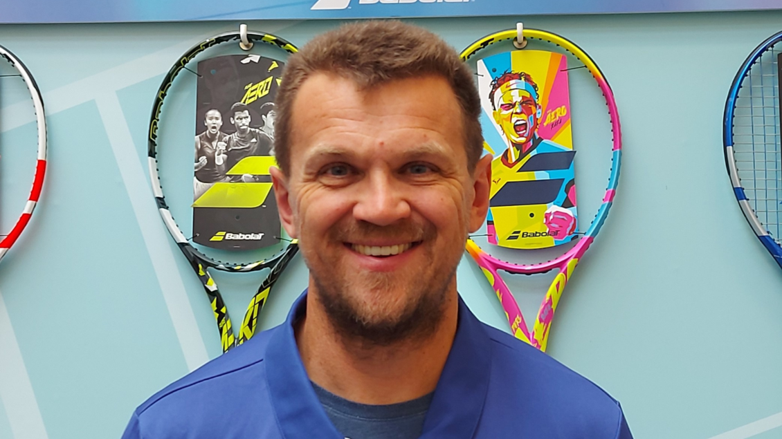 Ein lächelnder Mann trägt ein blaues Hemd und steht vor einer Wand mit Tennisschlägern. Im Hintergrund sind verschiedene Schläger und bunte Designs zu sehen. | © Mike Krug