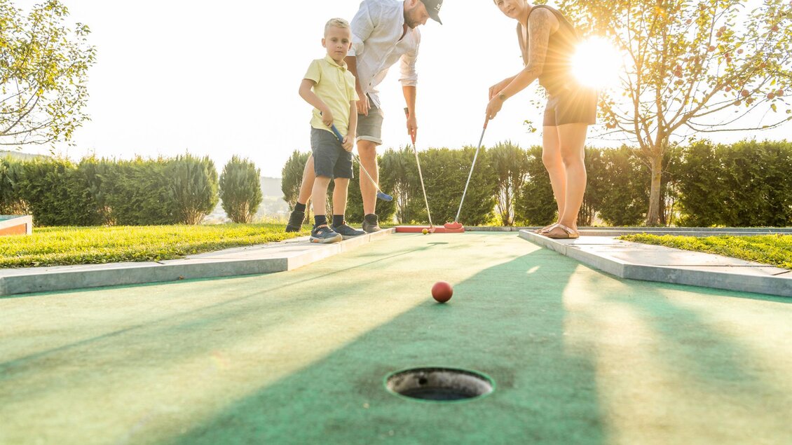 Eine Familie spielt Minigolf im Freien. Die Sonne scheint und es gibt grüne Bäume im Hintergrund. | © Lipizzanerheimat_Die Abbilderei