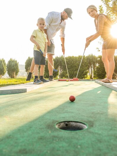 A family is playing mini-golf outdoors. The sun is shining and there are green trees in the background. | © Lipizzanerheimat_Die Abbilderei