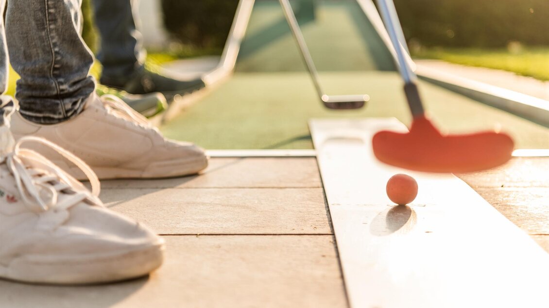 Eine Person spielt Minigolf auf einer Anlage. Der Golfball liegt auf dem Platz, während der Schläger bereit ist, ihn abzuschlagen. | © Lipizzanerheimat_Die Abbilderei