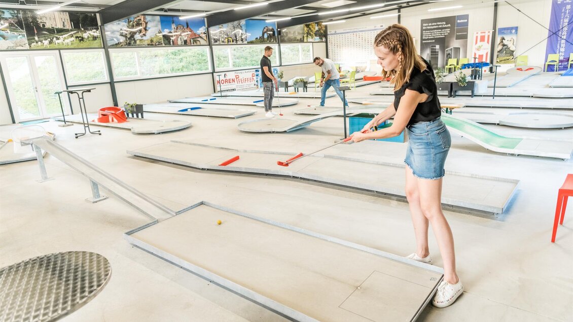 Eine Person spielt Minigolf in einer hellen, modernen Indoor-Anlage. Im Hintergrund sind weitere Spieler und verschiedene Minigolfbahnen zu sehen. | © Lipizzanerheimat_Die Abbilderei