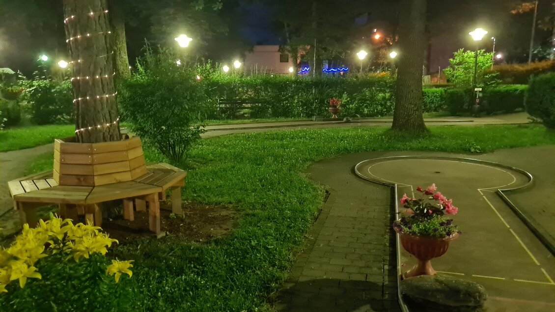 Ein ruhiger Park bei Nacht mit sanfter Beleuchtung. Eine Holzbank und bunte Blumen schmücken den Weg. | © Minigolfanlage Knittelfeld