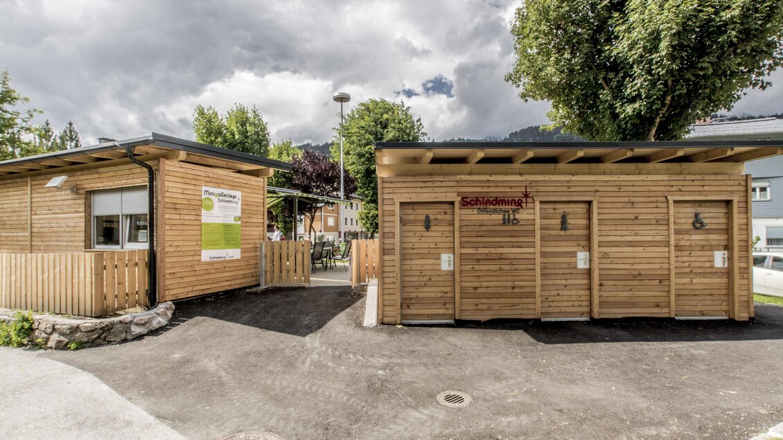 Zwei moderne Holzgebäude, umgeben von Bäumen und einem klaren Himmel. Das linke Gebäude hat eine Informationswand, während das rechte Gebäude mit mehreren Eingängen ausgestattet ist. | © Gerhard Pilz