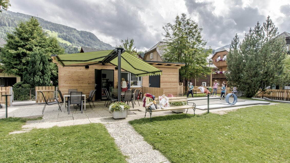 Ein gemütliches Holzhaus mit einem grünen Vordach in einer grünen Gartenlandschaft. Um das Haus herum befinden sich Stühle und Tische, und im Hintergrund sieht man einige Menschen und Bäume. | © Gerhard Pilz