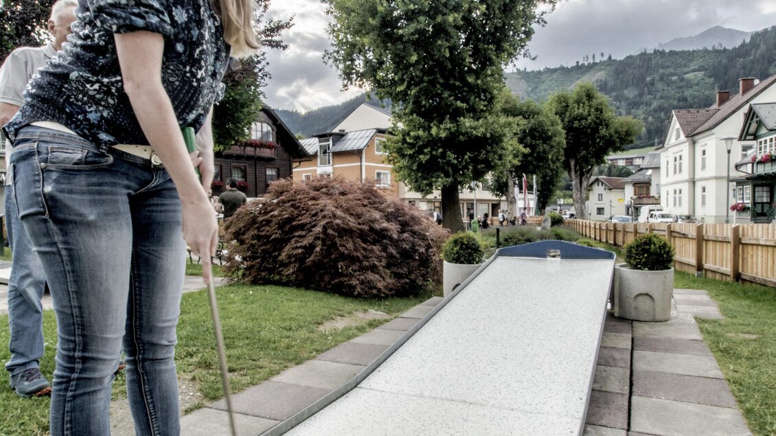 Eine Frau spielt Minigolf auf einer Bahn im Freien. Im Hintergrund sind Bäume und mehrere Häuser zu sehen. | © Gerhard Pilz