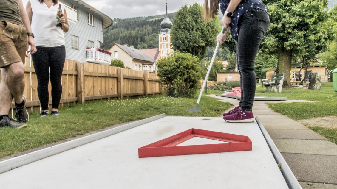 Eine Frau spielt Minigolf auf einem Outdoor-Kurs. Im Hintergrund stehen zwei Zuschauer und einige Bäume. | © Gerhard Pilz