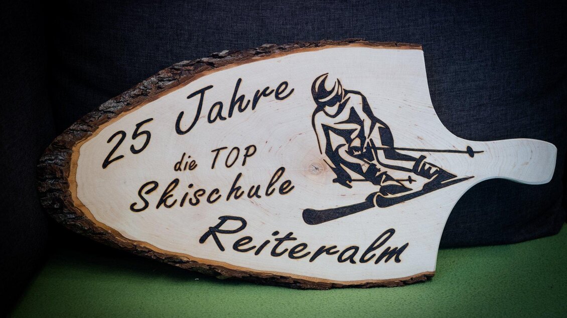 Ein handgefertigtes Schild feiert 25 Jahre die TOP Skischule Reiteralm. Es zeigt eine Skifahrer-Silhouette und hat eine rustikale Holzoptik. | © Minimi