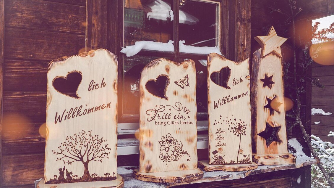 Eine Reihe von dekorativen Holzschildern mit verschiedenen Motiven und Schriftzügen steht vor einem Holzgebäude. Die Schilder sind mit einladenden Botschaften wie "Willkommen" und "Tritt ein" versehen. | © Minimi