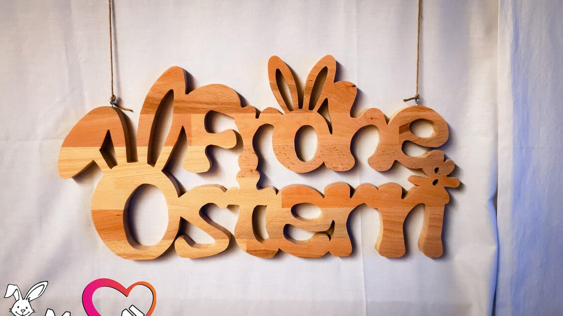Ein hölzernes Schild mit der Aufschrift „Frohe Ostern“ und dekorativen Hasenohren. Es hängt an zwei Schnüren und hat einen festlichen Charme.