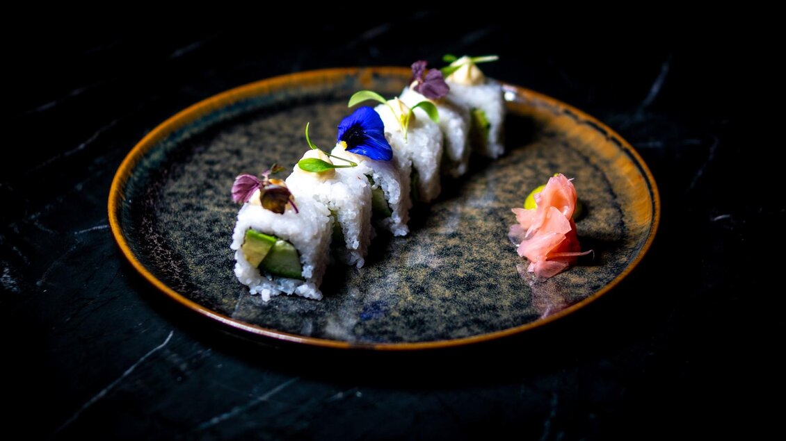 Ein elegantes Sushi-Gericht auf einem dunklen Teller. Die Röllchen sind mit essbaren Blumen und Ingwer dekoriert. | © Miss Cho - aiola