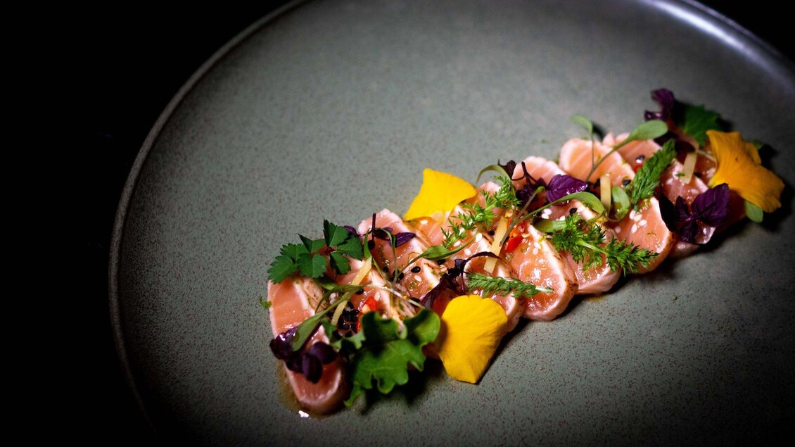 Ein elegantes Gericht mit dünn geschnittenem Fisch, garniert mit frischen Kräutern und essbaren Blüten. Die Präsentation ist kunstvoll auf einem schlichten Teller angeordnet. | © Miss Cho - aiola