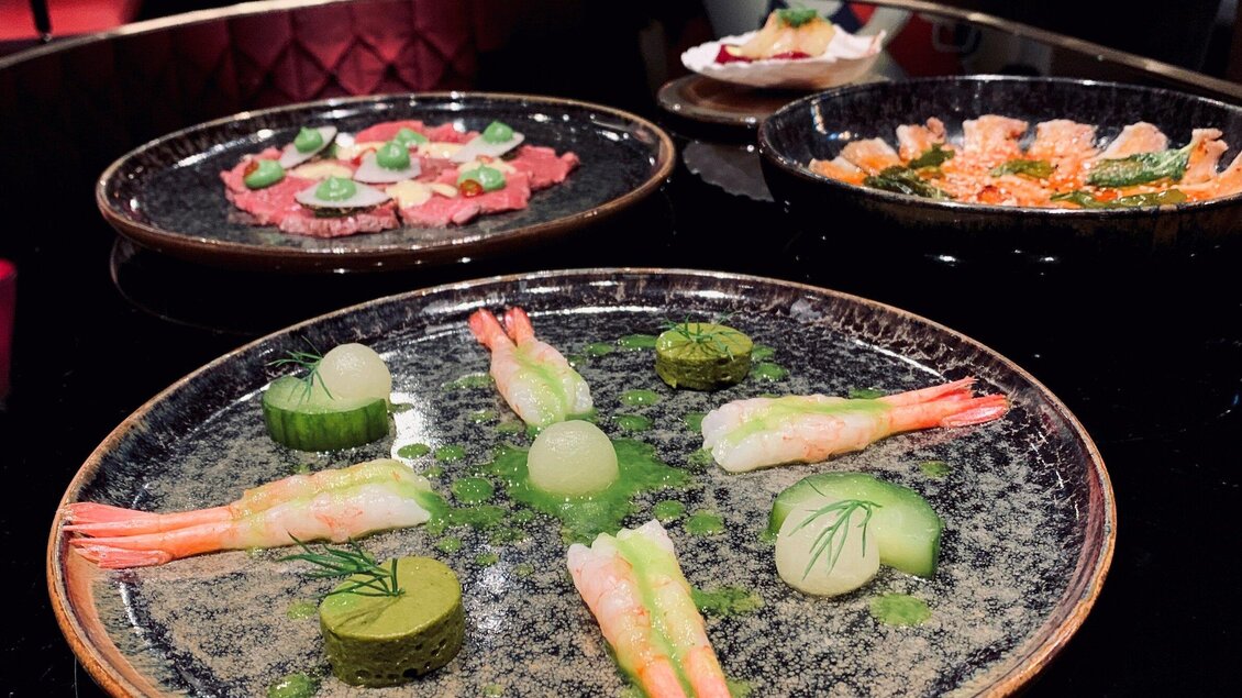 Ein elegantes Gericht mit Garnelen, Gurkenscheiben und grüner Sauce auf einem schwarzen Teller. Im Hintergrund sind weitere kunstvoll angerichtete Teller mit verschiedenen Speisen zu sehen. | © Miss Cho - aiola