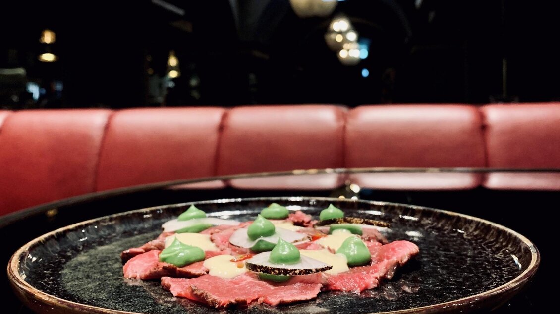 Ein kunstvoll angerichteter Teller mit Rindfleisch und grünen Saucenpunkten. Im Hintergrund sind stilvolle, gedämpfte Lichter und eine rote Sitzbank sichtbar. | © Miss Cho - aiola