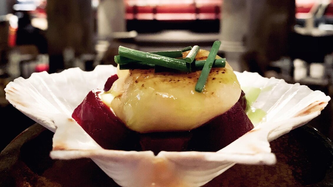 Ein elegant angerichtetes Gericht auf einer Muschelschale mit einer köstlichen Garnelenportion. Im Hintergrund ist eine stilvolle Restaurantatmosphäre zu sehen. | © Miss Cho - aiola