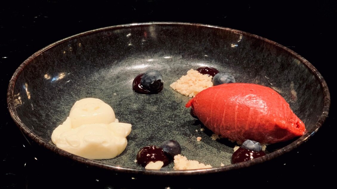 Ein kunstvoll angerichtetes Dessert in einer dunklen Schüssel. Es enthält ein rotes Sorbet, weiße Creme und verschiedene Beeren. | © Miss Cho - aiola