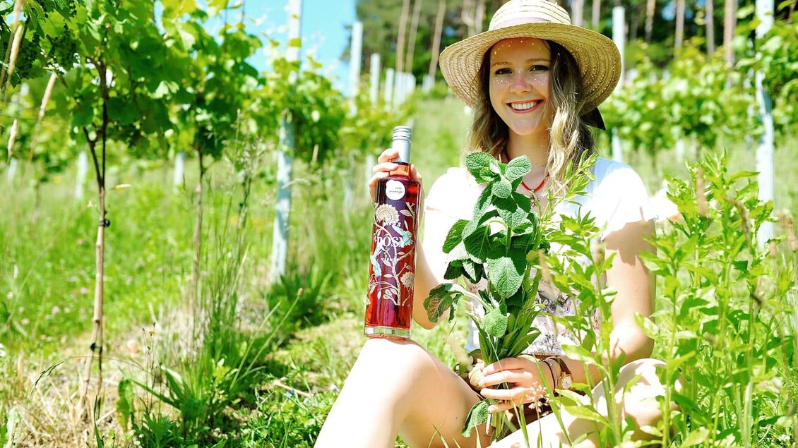 Eine lächelnde Frau sitzt in einem Weinberg und hält eine Flasche Roséwein sowie eine Pflanze in der Hand. Die Umgebung ist grün und sonnig, mit Weinreben im Hintergrund. | © Miss Rosy