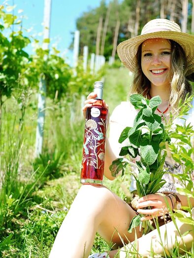 Eine lächelnde Frau sitzt in einem Weinberg und hält eine Flasche Roséwein sowie eine Pflanze in der Hand. Die Umgebung ist grün und sonnig, mit Weinreben im Hintergrund. | © Miss Rosy