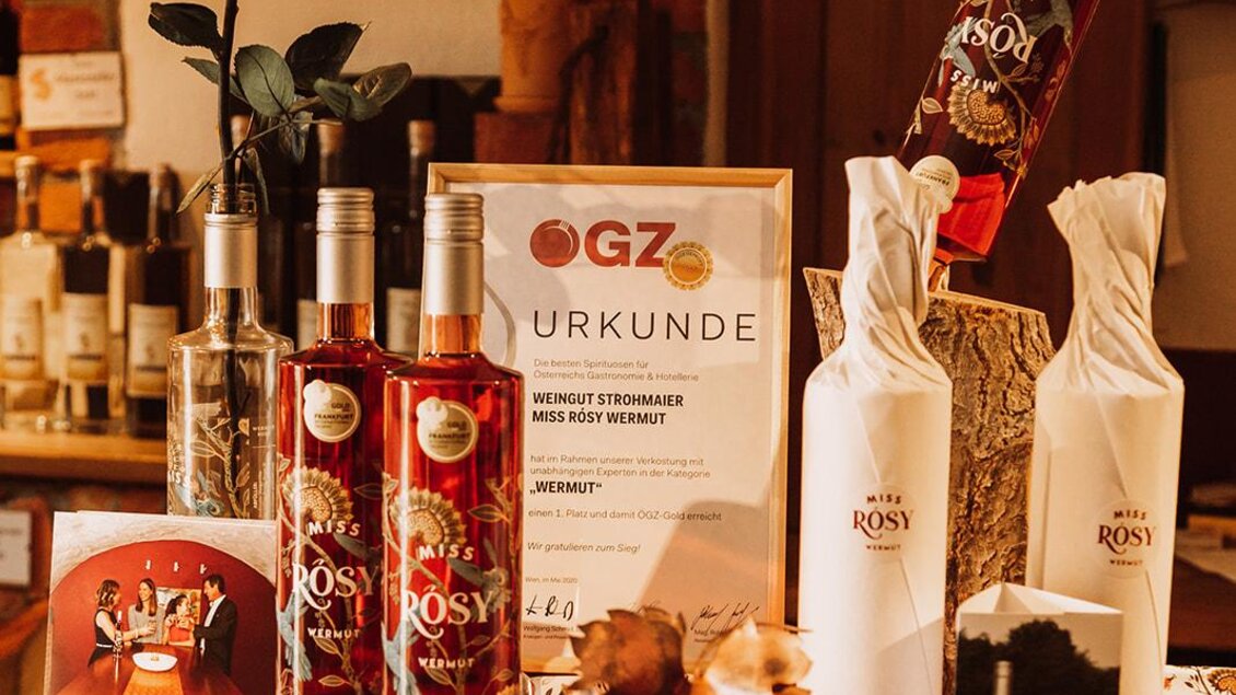 Eine stilvolle Präsentation von Flaschen und Produkten auf einem Holztisch. Im Hintergrund sind dekorative Elemente und eine Urkunde zu sehen. | © Miss Rosy