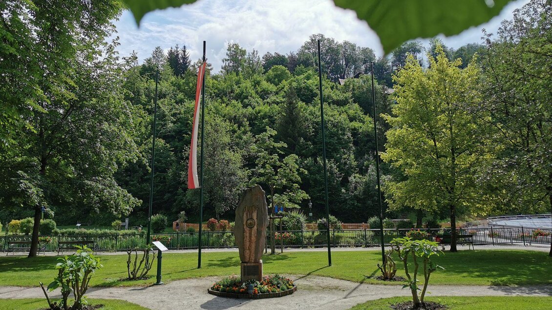Eine gepflegte Parkanlage mit grünen Bäumen und bunten Blumen. Im Mittelpunkt steht eine Statue umgeben von Flaggen und einem schönen Wald. | © TVB Ausseerland - Salzkammergut_Theresa Schwaiger