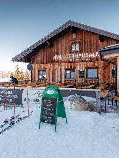 Eine Holzhütte im Schnee mit Sitzplätzen im Freien und Skiausrüstung vor der Tür. Im Hintergrund sind schneebedeckte Berge zu sehen. | © Mitterhausalm | Alpine Genusswelten