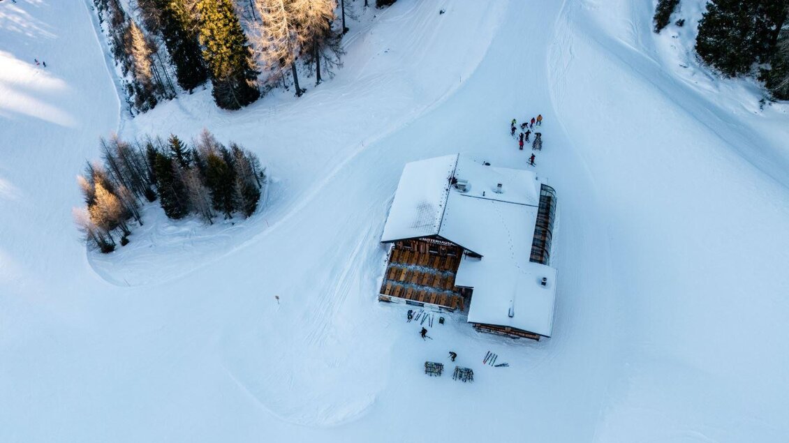 Ein gemütliches Holzhaus liegt inmitten einer verschneiten Landschaft. Umgeben von hohen Bäumen und einer Gruppe von Menschen, die im Schnee aktiv sind. | © Mitterhausalm | Alpine Genusswelten