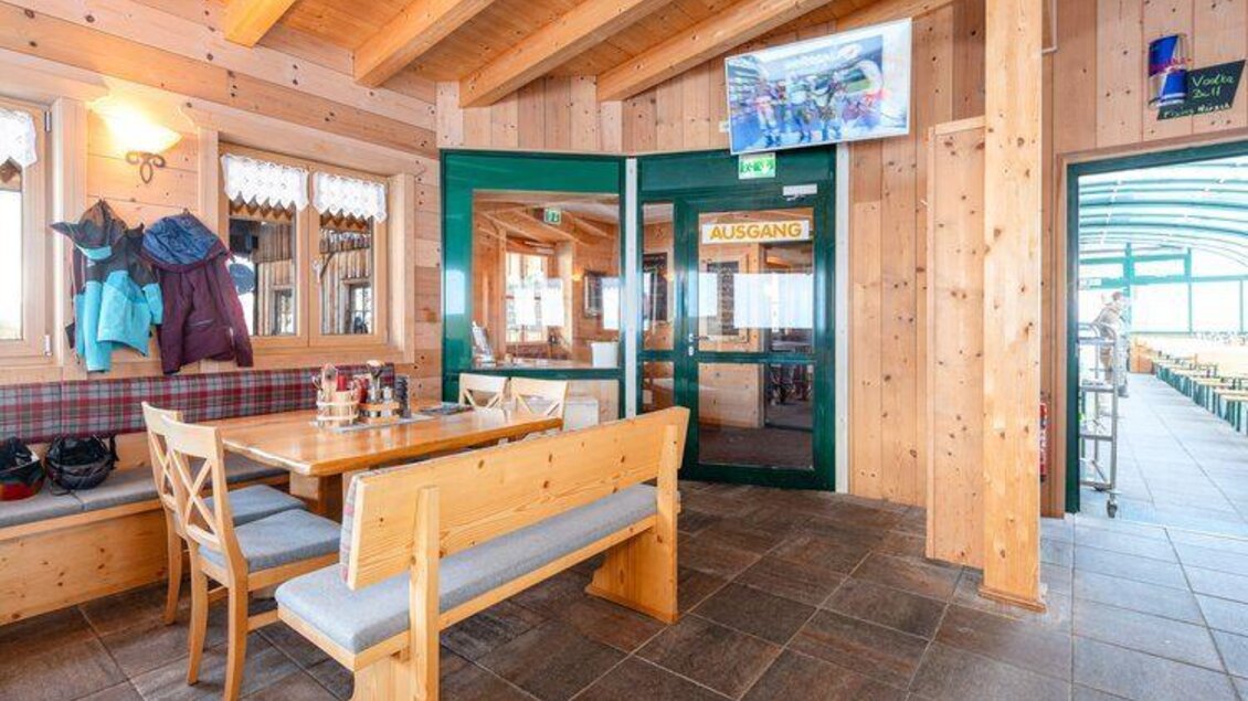 Ein rustikales Restaurant mit Holzverkleidung und einem großen Tisch. Durch ein Fenster sieht man den Ausgang und einen Blick nach draußen. | © Mitterhausalm | Alpine Genusswelten