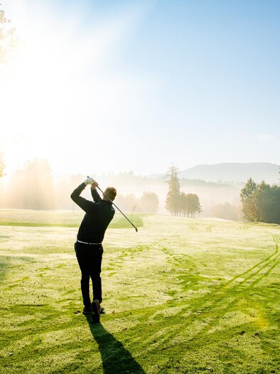 A golfer hits his ball on a foggy golf course. The sun gently shines through the trees, illuminating the green landscape. | © Region Graz - Mias Photoart