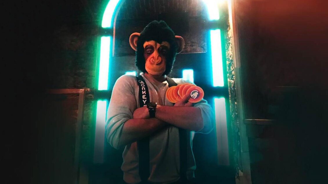 Person mit Affenmaske steht vor hellem Neonbogen und hält mehrere bunte runde Objekte in den Armen | © Monkeys