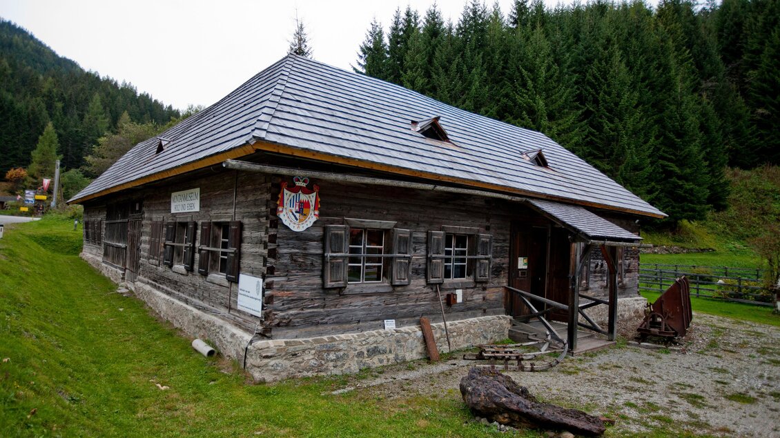 Ein traditionelles Holzhaus mit einem grauen Dach in einer grünen Landschaft. Im Hintergrund sind Nadelbäume zu sehen. | © Holzwelt Murau