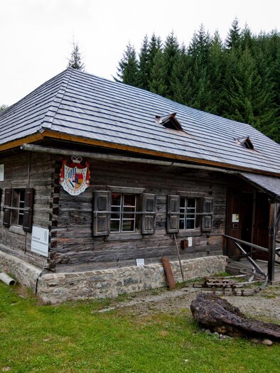A traditional wooden house with a gray roof in a green landscape. In the background, coniferous trees are visible. | © Holzwelt Murau